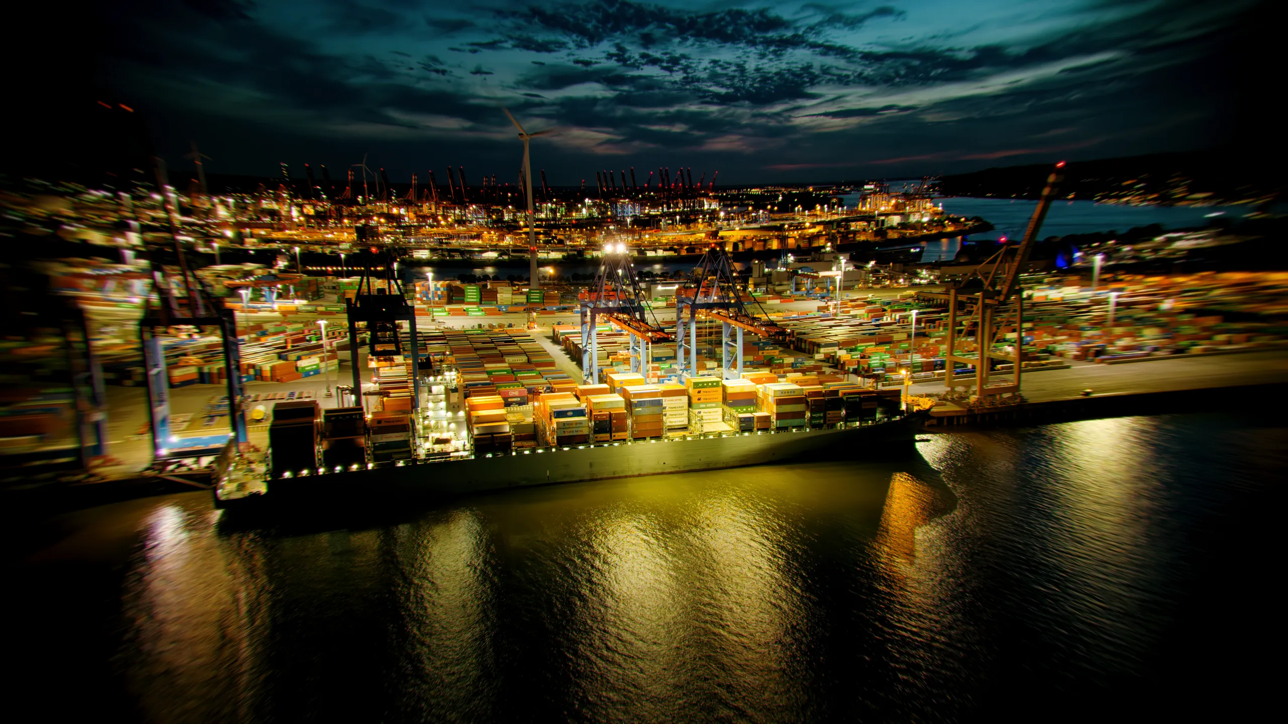 Luftaufnahme eines beleuchteten Containerterminals im Hamburger Hafen bei Nacht mit Containerbrücken, Frachtschiff und dramatischem Himmel.