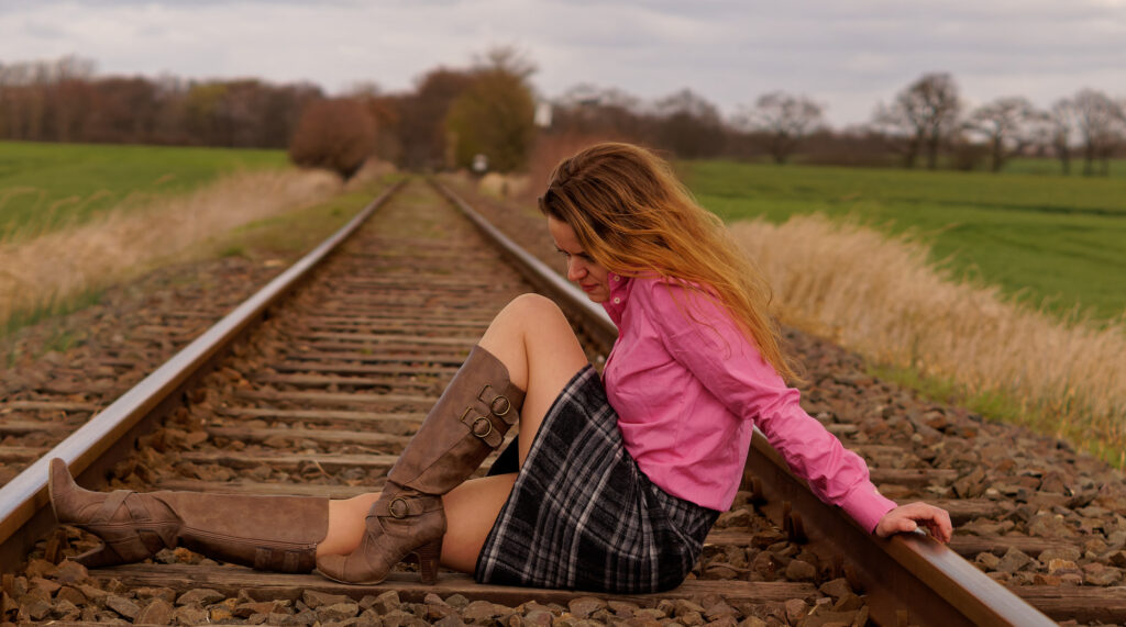 unge Frau mit langen, hellbraunen Haaren, pinkfarbener Bluse und kariertem Rock sitzt auf Eisenbahnschienen inmitten einer grünen Landschaft.
