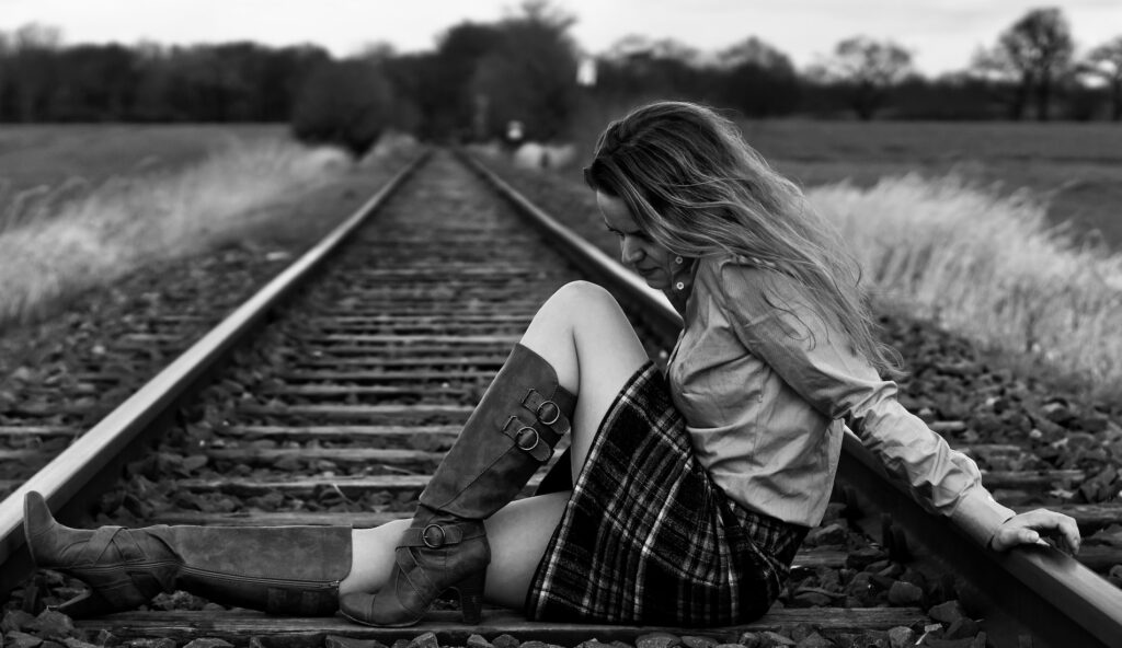 Schwarz-Weiß-Porträt einer Frau mit langen Haaren, die nachdenklich zwischen Eisenbahnschienen sitzt. Die Gleise verlaufen durch eine weite Landschaft bis zum Horizont.