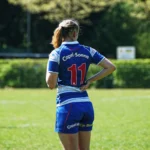 Rugbyspielerin von Bayern München mit blauem Trikot und der Rückennummer 11 steht mit einer Hand an der Hüfte auf dem Spielfeld.