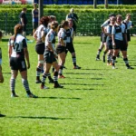 Rugbyspielerinnen des FC St. Pauli in braun-weißen Trikots bereiten sich auf einen ruhenden Kick gegen Bayern München vor, während der Rugbyball auf dem Rasen steht.