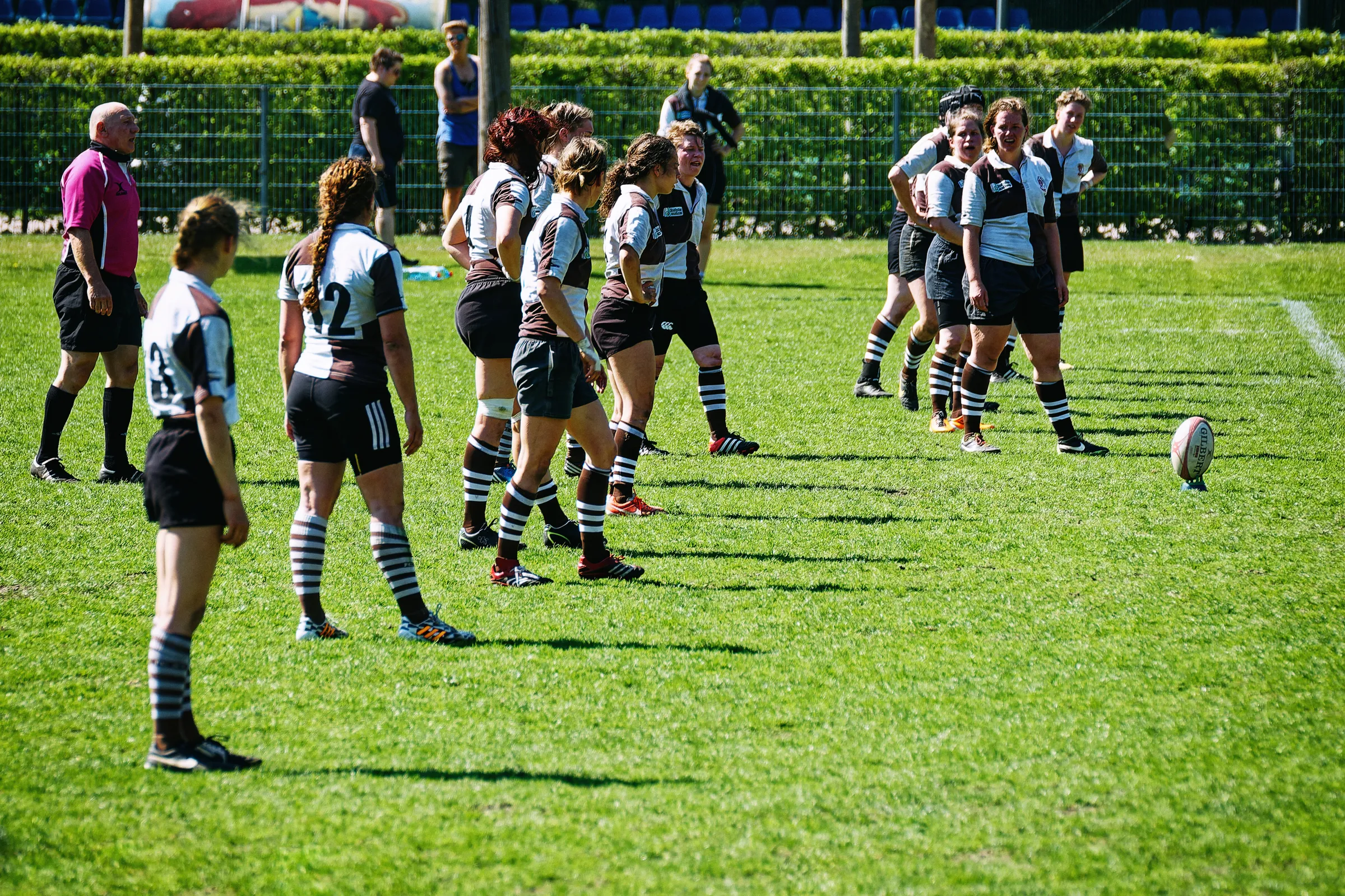 Rugbyspielerinnen des FC St. Pauli in braun-weißen Trikots bereiten sich auf einen ruhenden Kick gegen Bayern München vor, während der Rugbyball auf dem Rasen steht.
