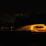 Brennende Stahlwolle erzeugt bei Nacht einen goldenen Funkenkreis mit weit auslaufenden Lichtspuren vor dunkler Baumkulisse.
