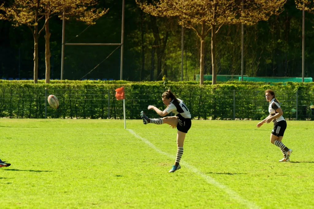 Rugbyspielerin des FC St. Pauli im braun-weißen Trikot tritt den Rugbyball mit ausgestrecktem Bein über das Spielfeld im Spiel gegen Bayern München.