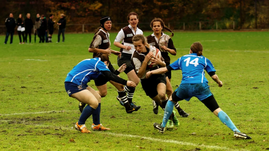Damenrugby-Spielerin des FC St. Pauli im braun-weißen Trikot trägt den Rugbyball durch die Abwehr von Bayern München, während zwei Spielerinnen in Blau versuchen, sie zu stoppen.