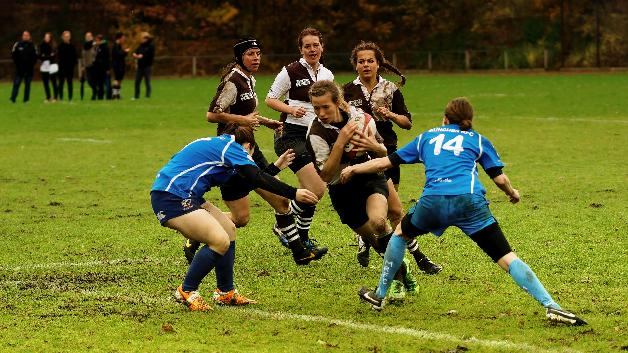 Damenrugby-Spielerin des FC St. Pauli im braun-weißen Trikot trägt den Rugbyball durch die Abwehr von Bayern München, während zwei Spielerinnen in Blau versuchen, sie zu stoppen.