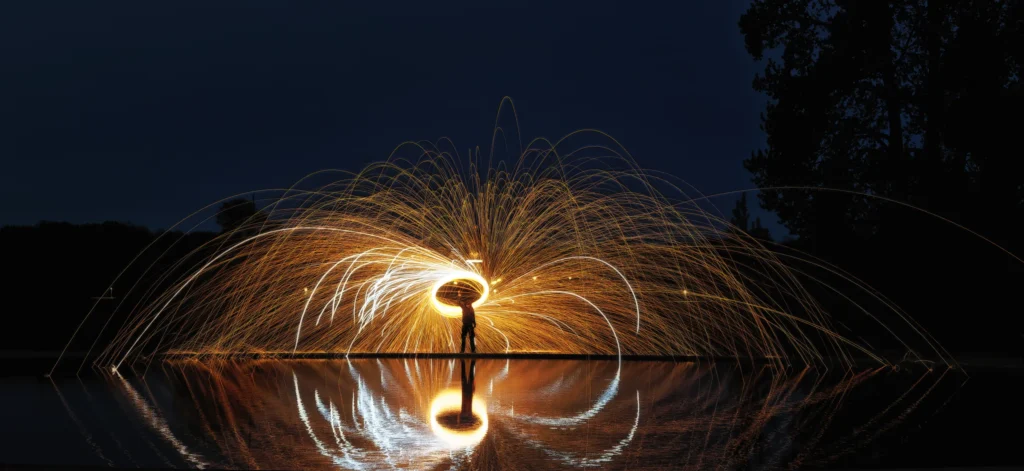 Goldener Funkenregen aus brennender Stahlwolle bildet einen leuchtenden Kreis, dessen Licht sich eindrucksvoll im Wasser spiegelt.