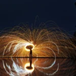 Goldener Funkenregen aus brennender Stahlwolle bildet einen leuchtenden Kreis, dessen Licht sich eindrucksvoll im Wasser spiegelt.