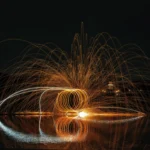 Brennende Stahlwolle zeichnet spiralförmige Lichtkreise und einen weit gespannten Funkenregen über einer spiegelnden Wasserfläche.