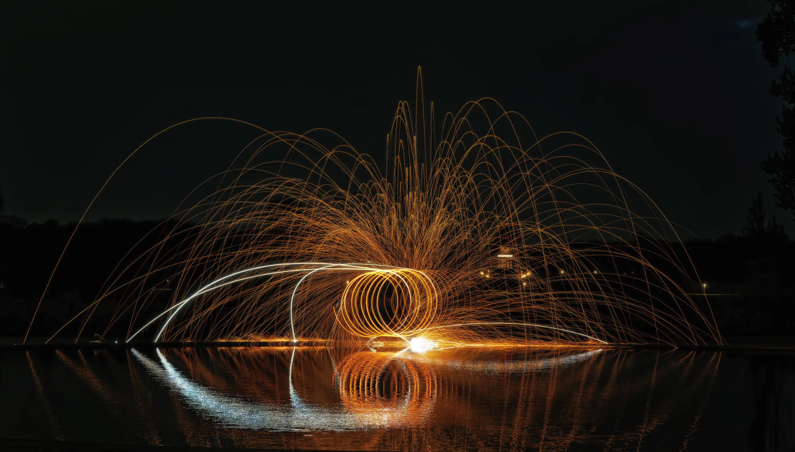Brennende Stahlwolle zeichnet spiralförmige Lichtkreise und einen weit gespannten Funkenregen über einer spiegelnden Wasserfläche.