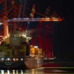 Heck des Containerschiffs MSC Bhavya V am Hamburger Hafen unter großen beleuchteten Containerbrücken bei Nacht.