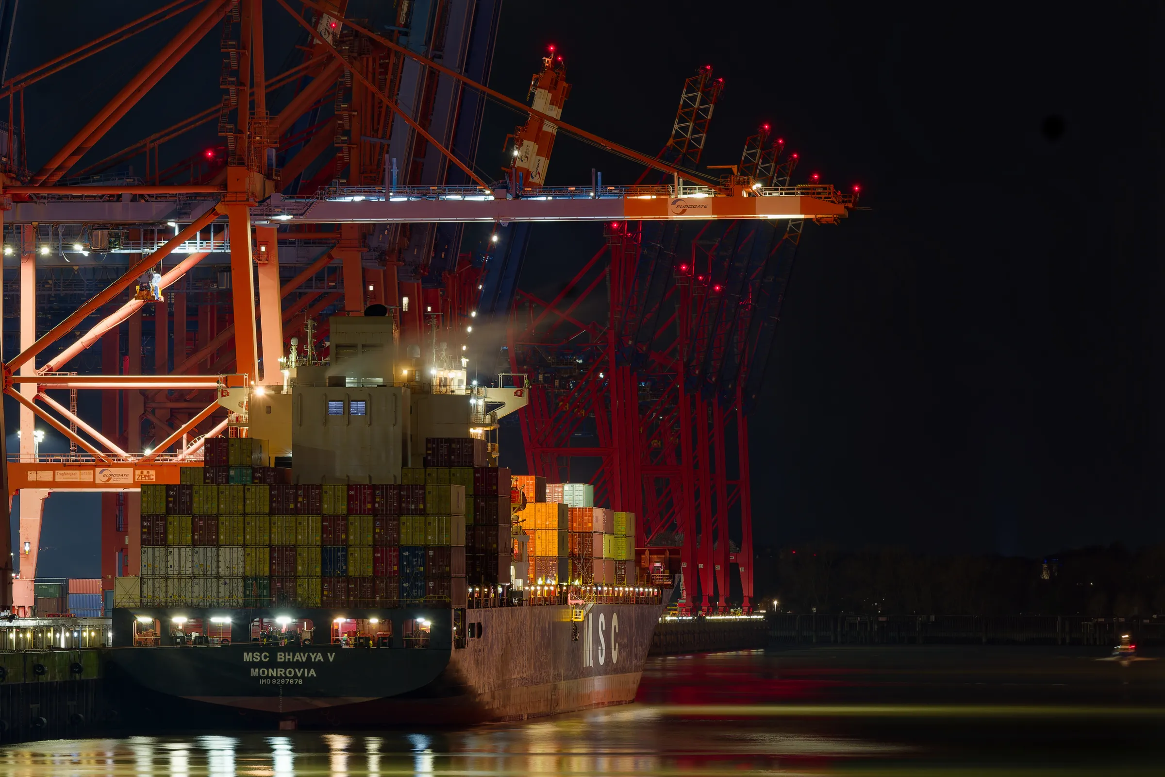 Heck des Containerschiffs MSC Bhavya V am Hamburger Hafen unter großen beleuchteten Containerbrücken bei Nacht.