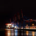 Nächtlicher Blick entlang der beleuchteten Containerbrücken am Terminal Burchardkai mit Containerschiff und Lichtspiegelungen auf der Elbe.