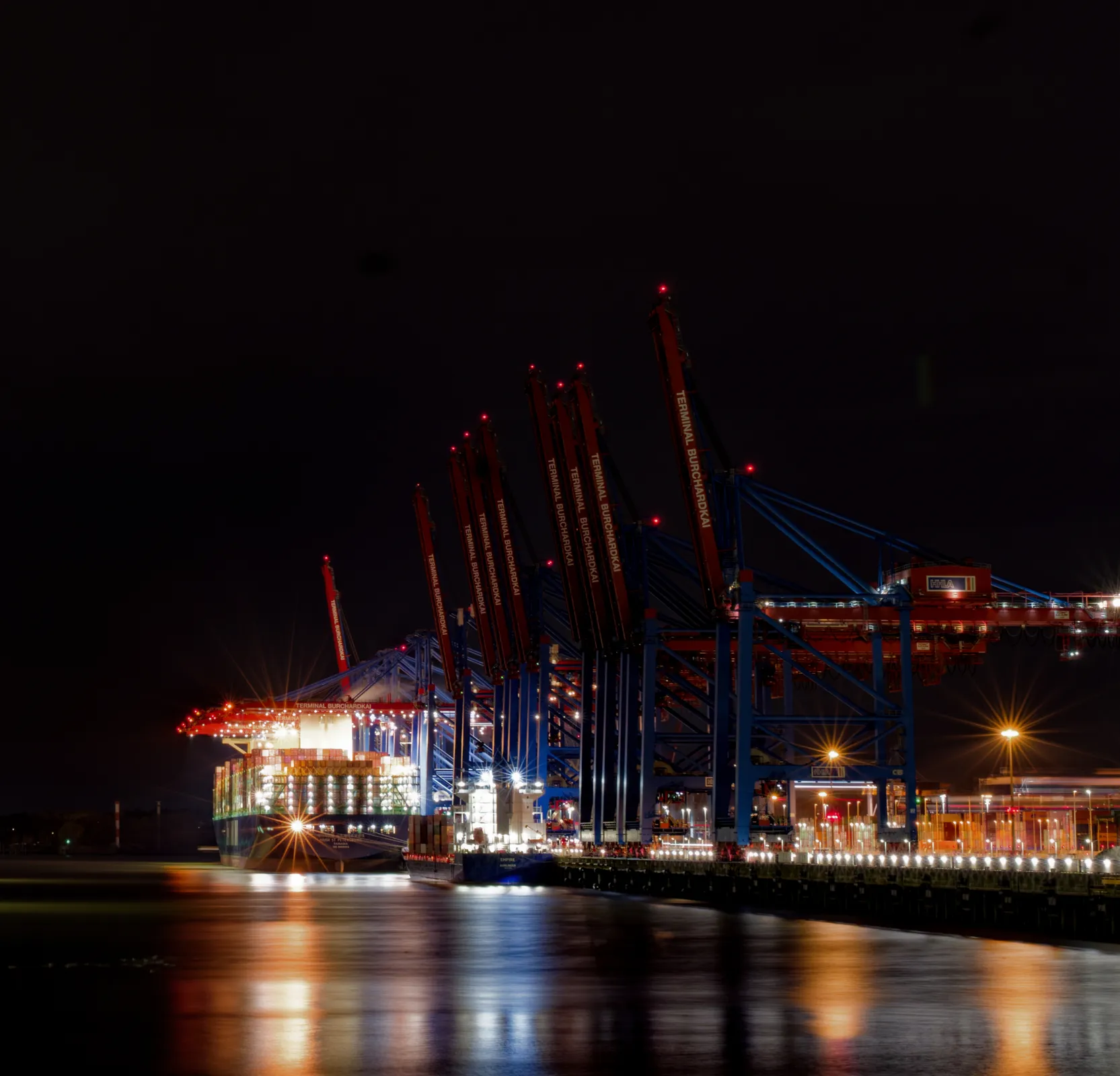 Nächtlicher Blick entlang der beleuchteten Containerbrücken am Terminal Burchardkai mit Containerschiff und Lichtspiegelungen auf der Elbe.