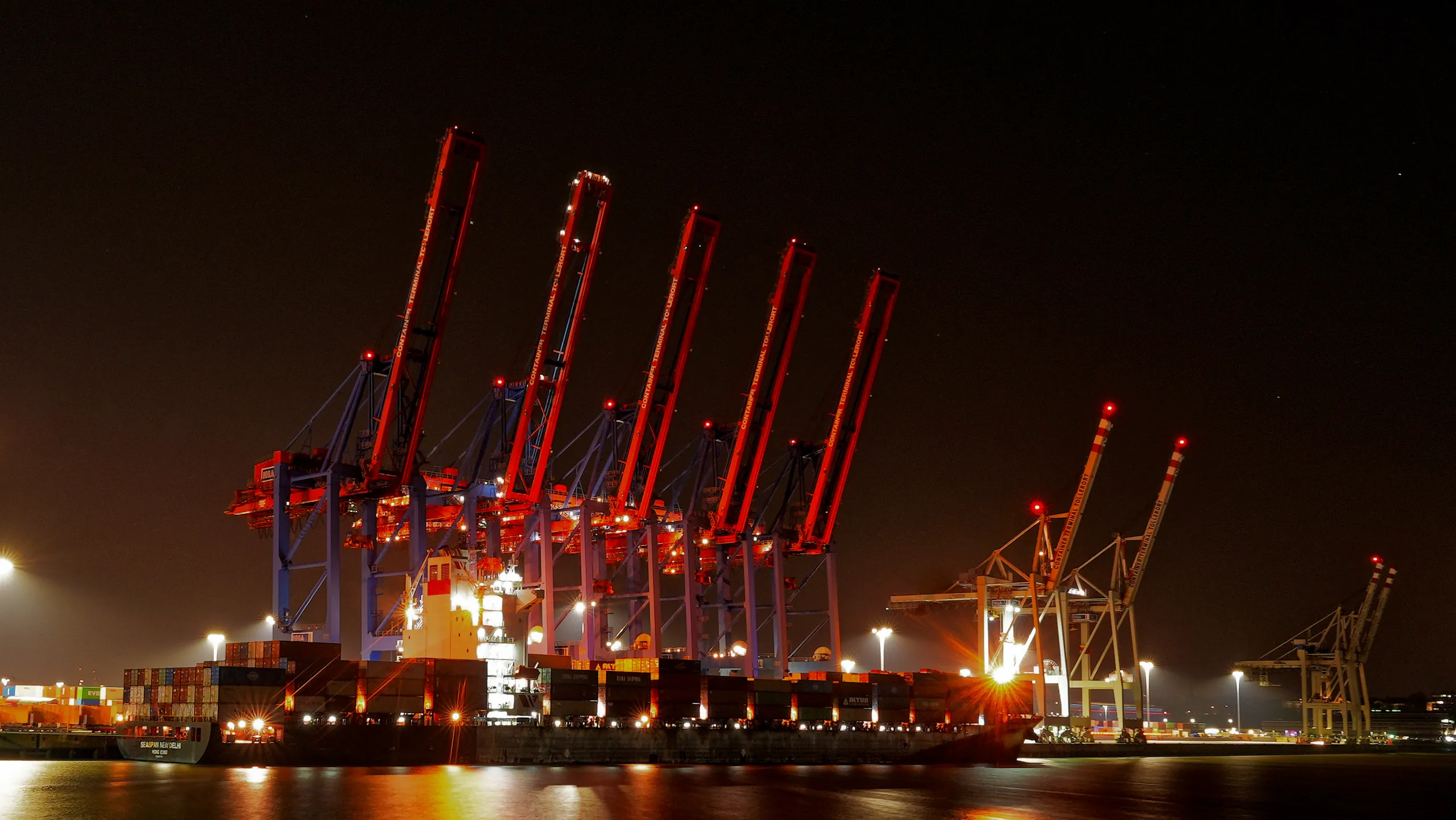 Rot beleuchtete Containerbrücken und ein großes Containerschiff am Containerterminal Altenwerder im Hamburger Hafen bei Nacht.