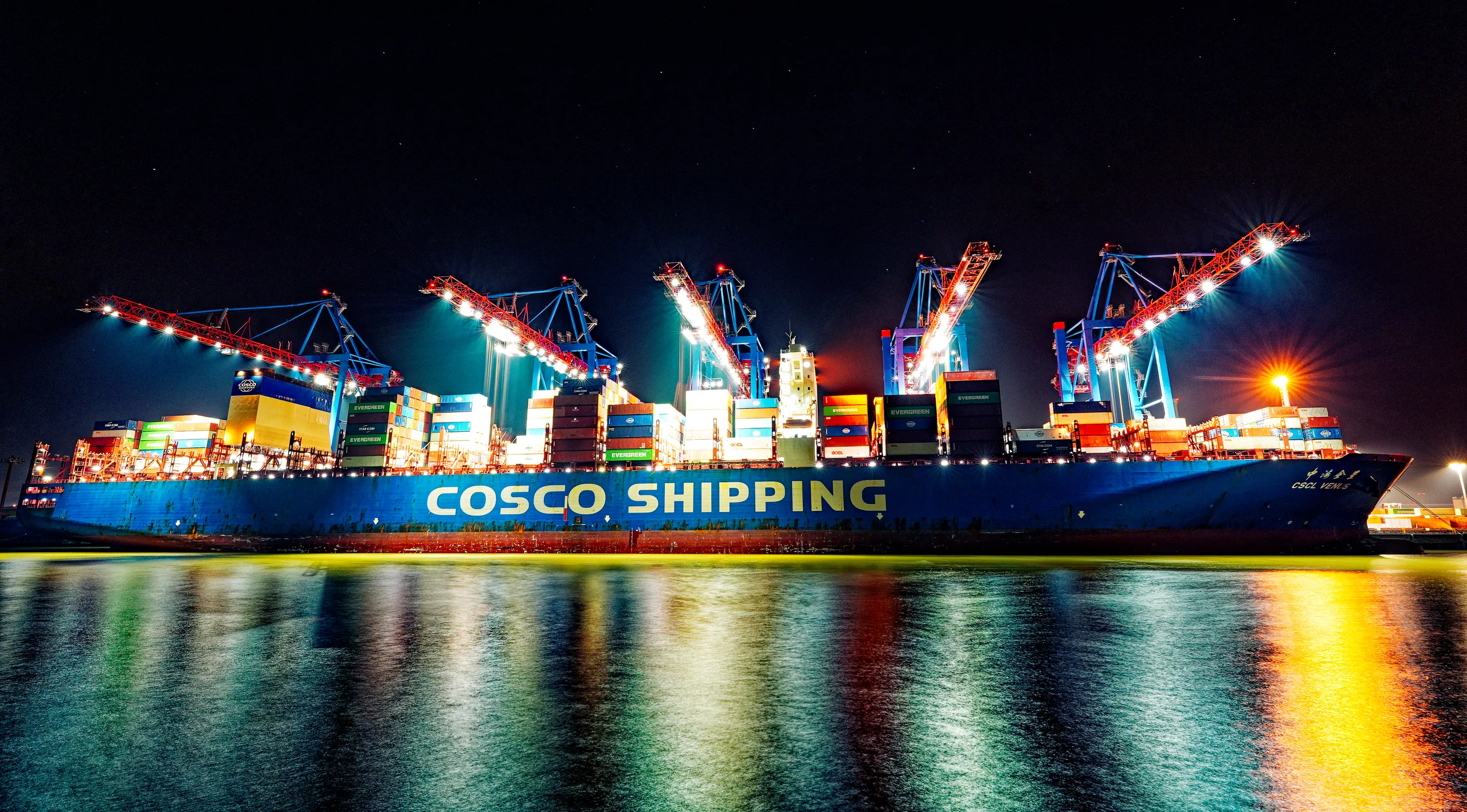 Großes blaues Containerschiff von COSCO Shipping bei nächtlichen Verladearbeiten unter beleuchteten Containerbrücken im Hamburger Hafen.