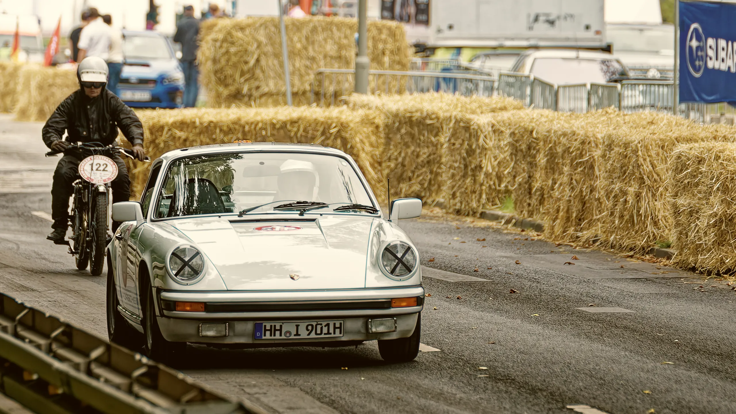 Silberner klassischer Porsche 911 G-Modell auf der abgesperrten Rennstrecke, gefolgt von einem historischen Motorradfahrer.