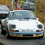 Weißer klassischer Porsche 911 mit blauen Rennstreifen und abgeklebten Rundscheinwerfern vor einem historischen Volkswagen auf der Rennstrecke. Weißer klassischer Porsche 911 mit blauen Rennstreifen und abgeklebten Rundscheinwerfern vor einem historischen Volkswagen auf der Rennstrecke. Die Karosserie erinnert an einen frühen 911 im historischen Rennsportstil. Eine genaue Untervariante lässt sich aus der Frontansicht nicht sicher bestimmen.