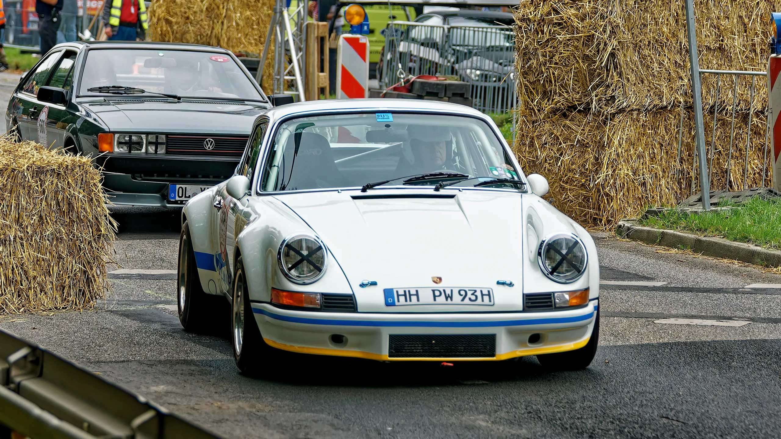 Weißer klassischer Porsche 911 mit blauen Rennstreifen und abgeklebten Rundscheinwerfern vor einem historischen Volkswagen auf der Rennstrecke. Die Karosserie erinnert an einen frühen 911 im historischen Rennsportstil. Eine genaue Untervariante lässt sich aus der Frontansicht nicht sicher bestimmen.