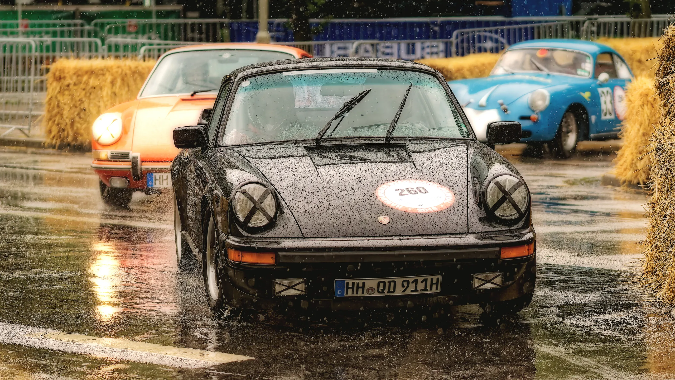 Schwarzer Porsche 911 G-Modell mit Startnummer 260 auf nasser Rennstrecke, gefolgt von einem orangefarbenen Porsche 911 und einem blauen Porsche 356.