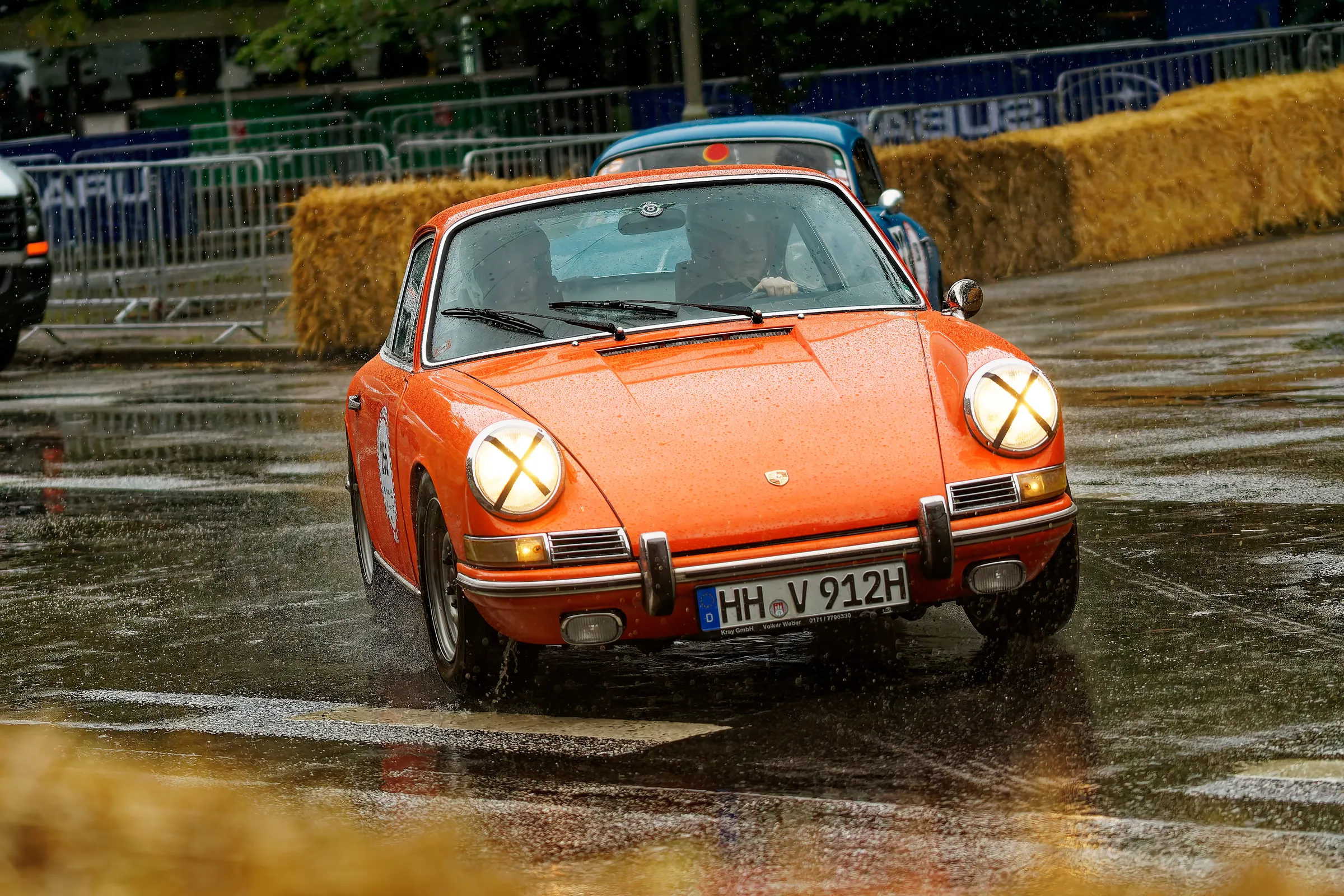 Orangefarbener klassischer Porsche 911 mit abgeklebten Scheinwerfern bei starkem Regen auf der historischen Rennstrecke in Hamburg.