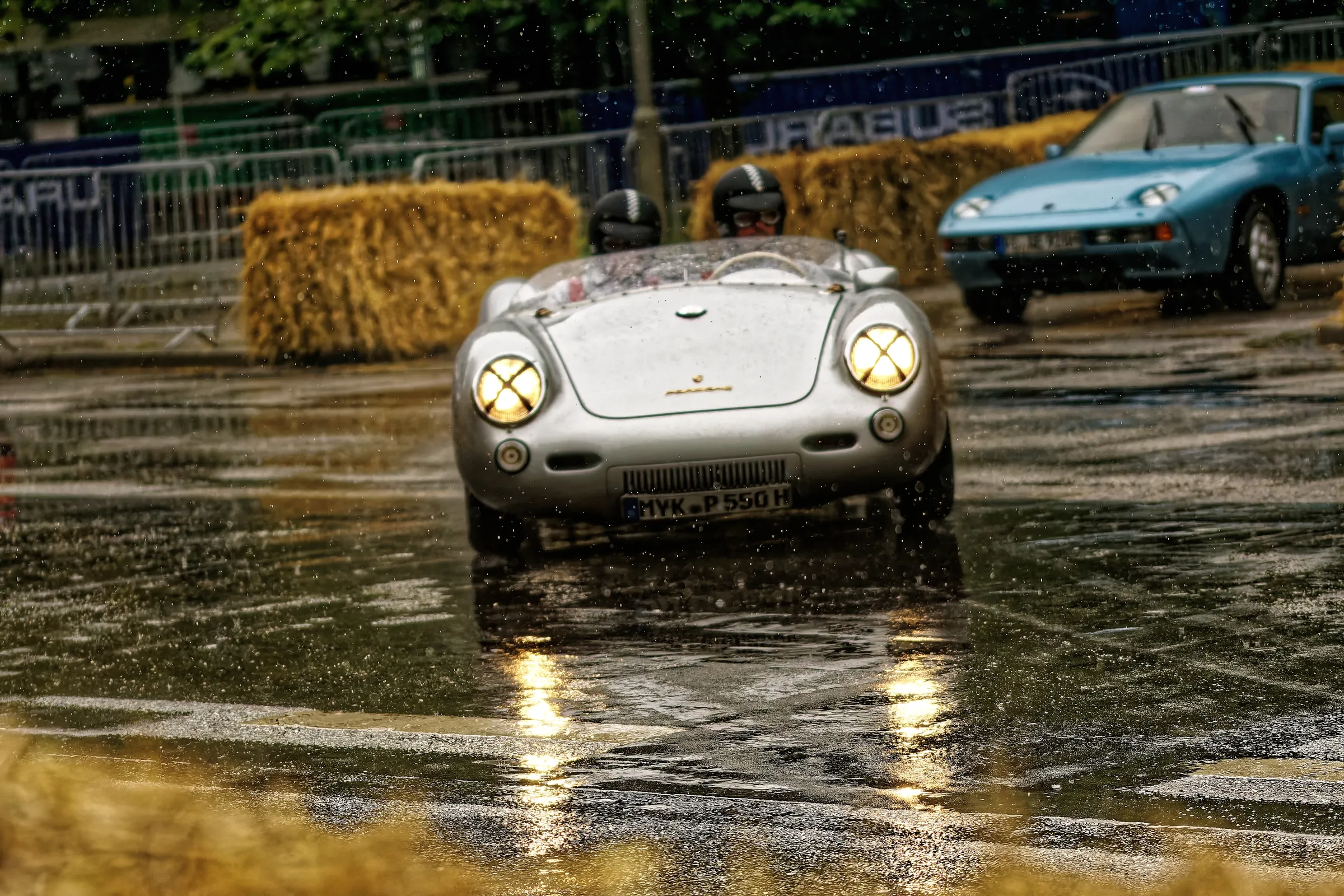 Silberner Porsche 550 Spyder mit zwei behelmten Insassen und eingeschalteten Rundscheinwerfern auf einer regennassen Rennstrecke.