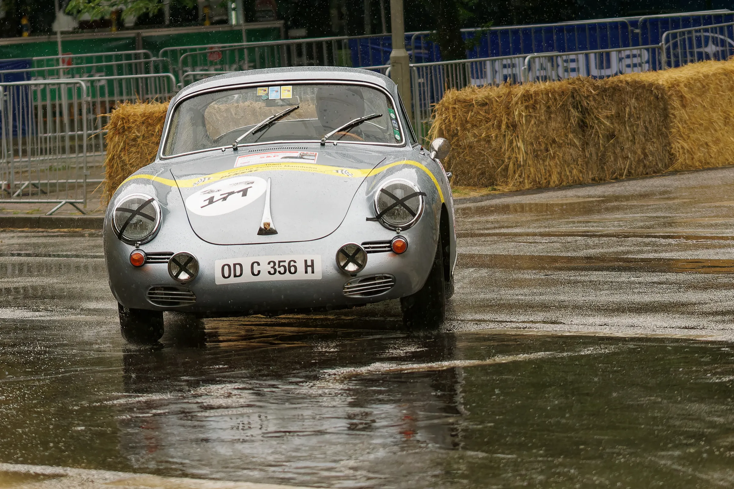 Silberner Porsche 356 Coupé mit Startnummer 144 und abgeklebten Rundscheinwerfern auf regennasser Fahrbahn.