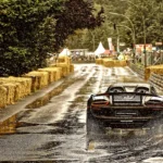 Grauer Porsche 918 Spyder mit großem Heckflügel auf regennasser Fahrbahn Weißer Porsche 911 GT3 RS mit großem Heckflügel und eingeschalteten Scheinwerfern auf regennasser Straße.