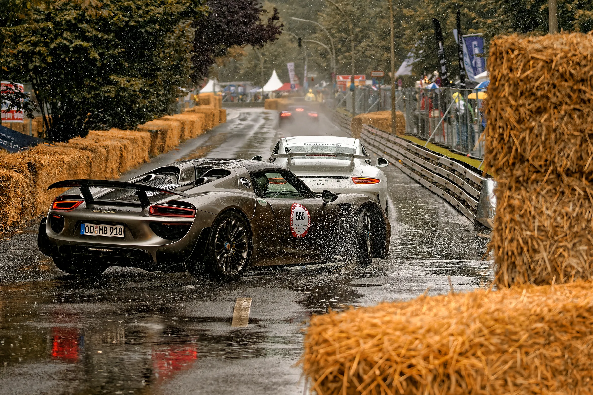 Grauer Porsche 918 Spyder mit großem Heckflügel auf regennasser Fahrbahn, dahinter ein weißer Porsche 911 GT3.