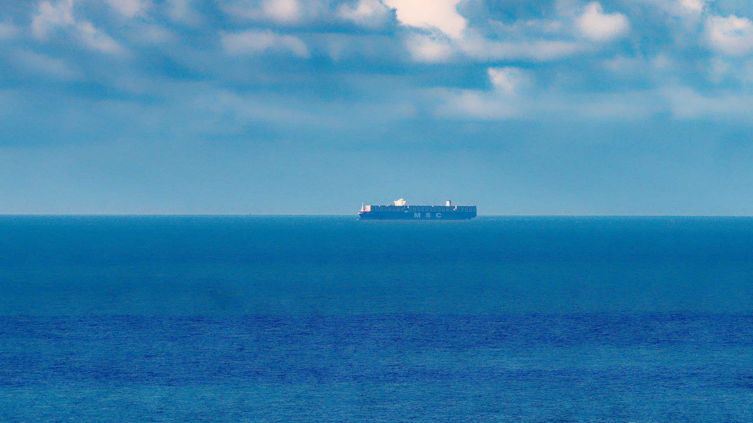 Containerschiff der Reederei MSC am Horizont auf dem offenen Meer unter blauem Himmel mit vereinzelten Wolken.