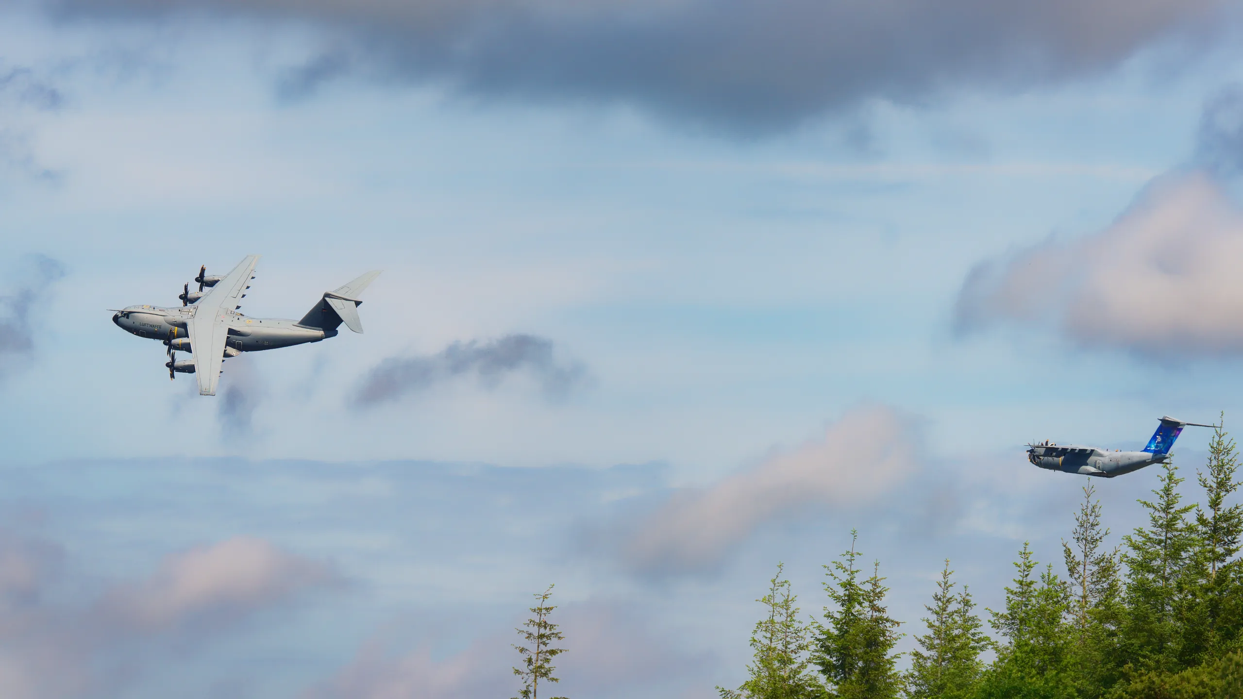 Zwei militärische Transportflugzeuge mit Propellertriebwerken im gemeinsamen Flug vor blauem Himmel und aufgelockerten Wolken.