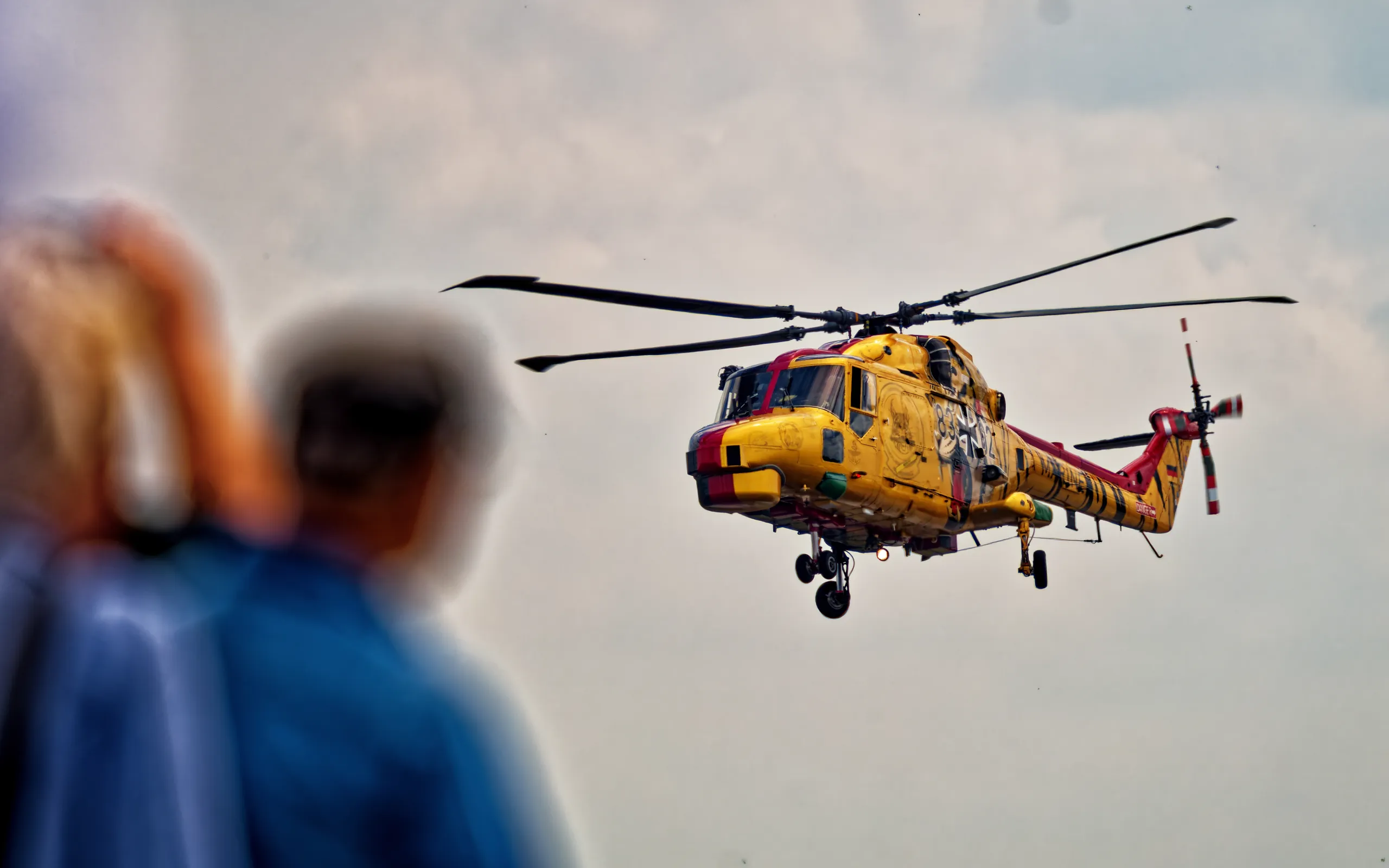 Gelb-roter Sea-Lynx-Hubschrauber im Flug über Nordholz, eingerahmt von unscharfen Zuschauern im Vordergrund.