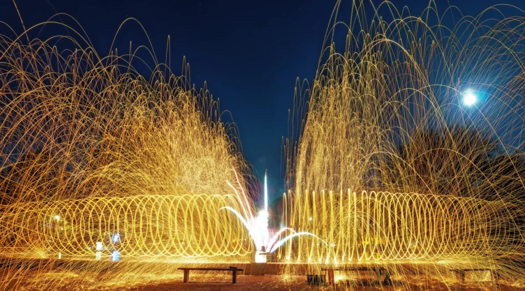 Goldene Funken aus brennender Stahlwolle bilden zwei große spiralförmige Lichtformationen und einen dichten Funkenregen vor dunklem Nachthimmel