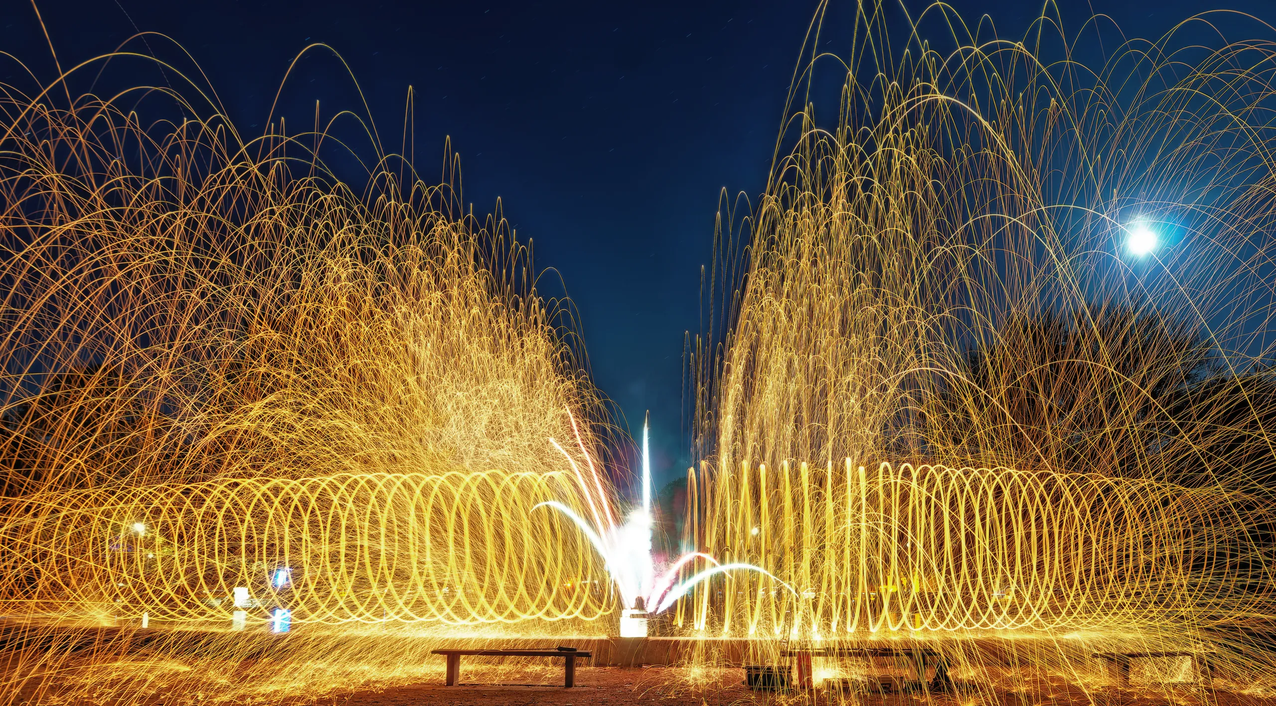 Goldene Funken aus brennender Stahlwolle bilden zwei große spiralförmige Lichtformationen und einen dichten Funkenregen vor dunklem Nachthimmel