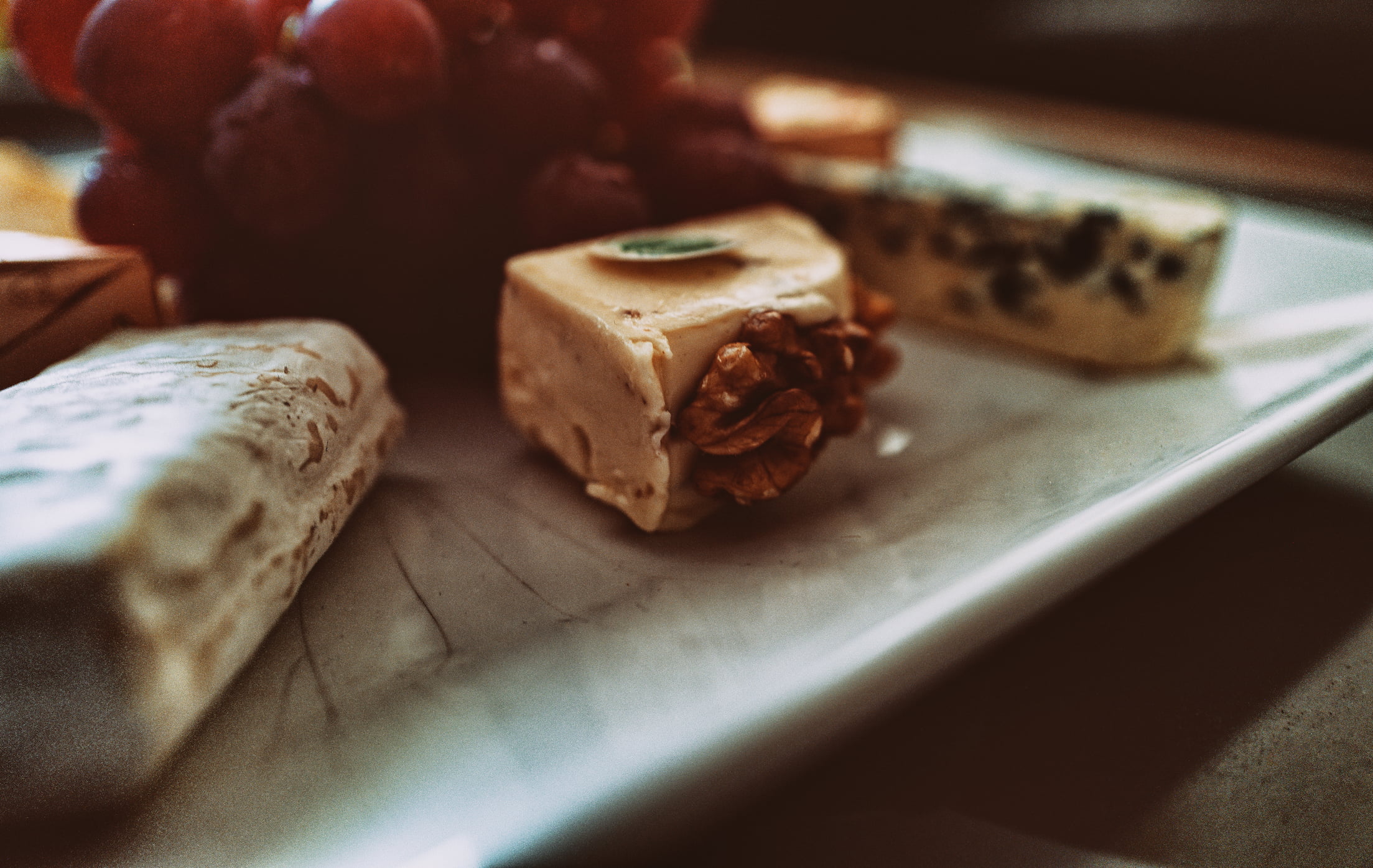 Nahaufnahme einer Käseauswahl mit cremigem Haselnusskäse, Walnüssen und Weintrauben auf einer weißen Servierplatte