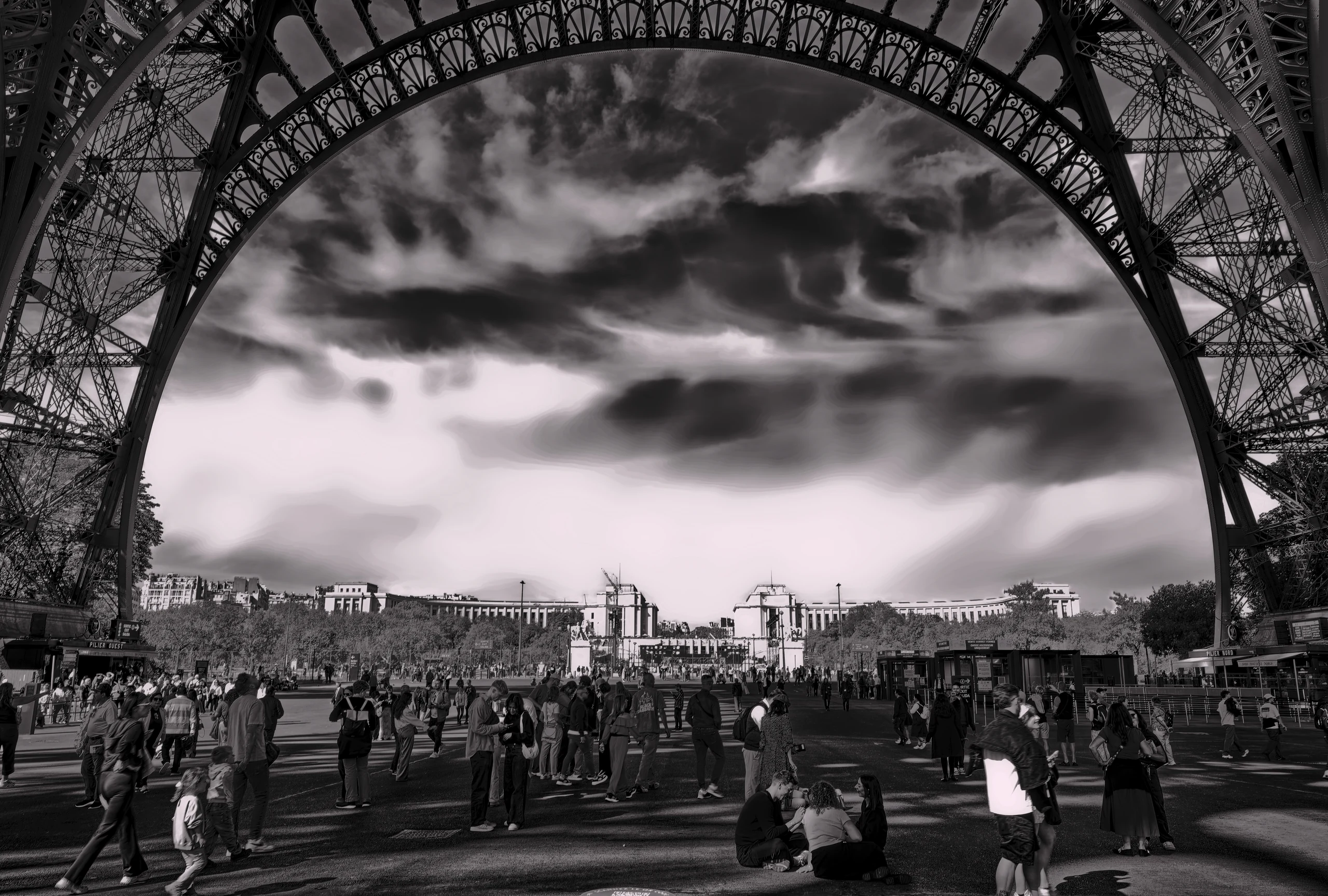 Schwarz-Weiß-Fotografie unter dem Eiffelturm mit Blick auf den Platz und dramatische Wolken